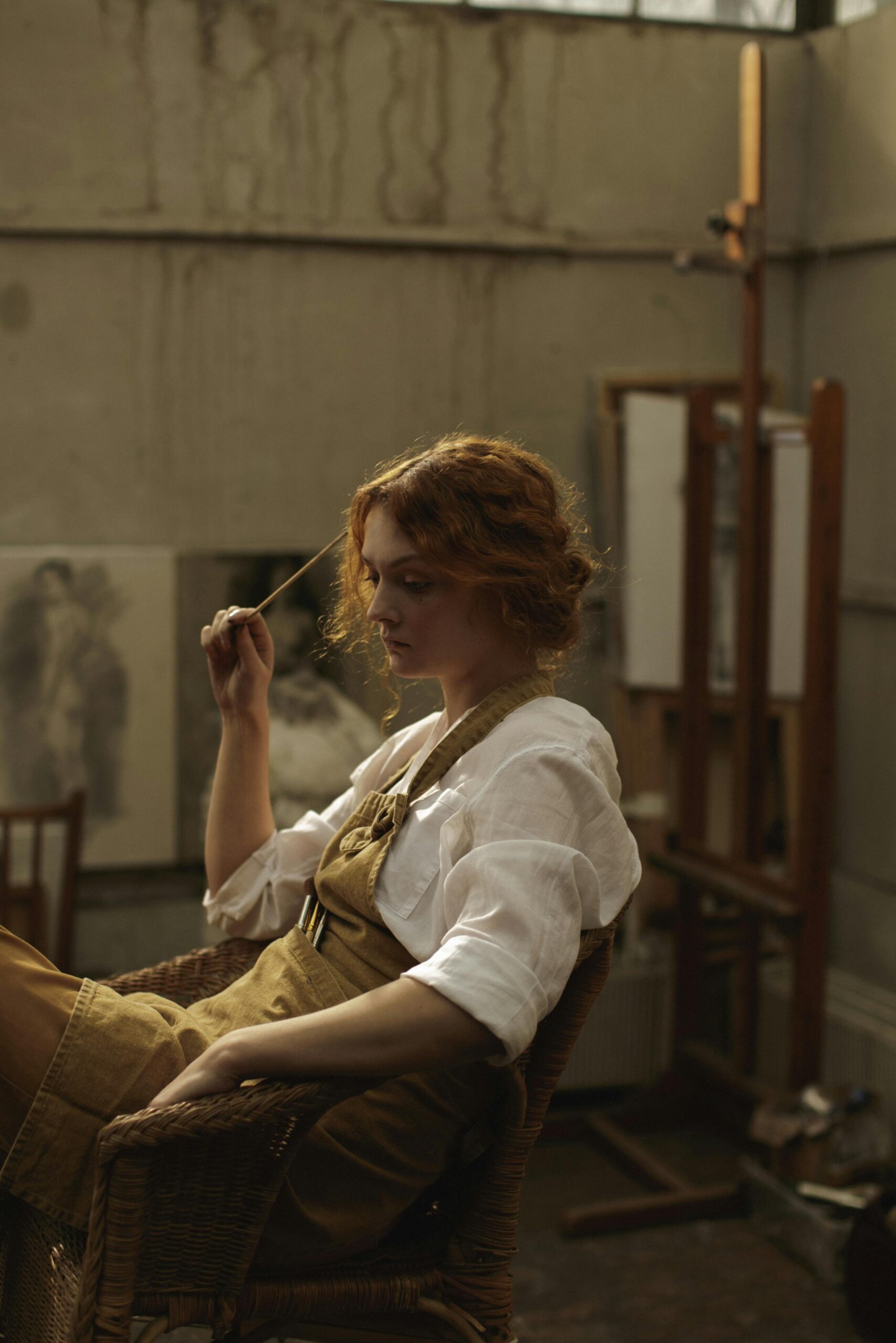 Young female artist in deep thought, sitting in a rustic studio. Shows creativity and contemplation.